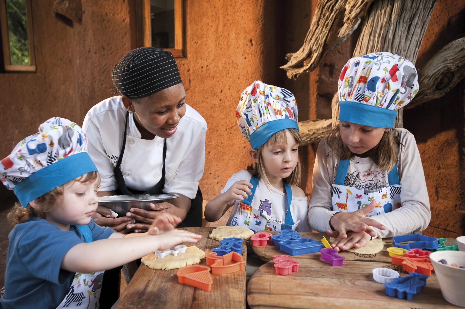 10-Madikwe-Safari-Lodge-malaria-free-safari-Baking A group of young children wearing colourful chef hats and aprons excitedly learn baking skills from a smiling chef, working together at a wooden table outdoors.