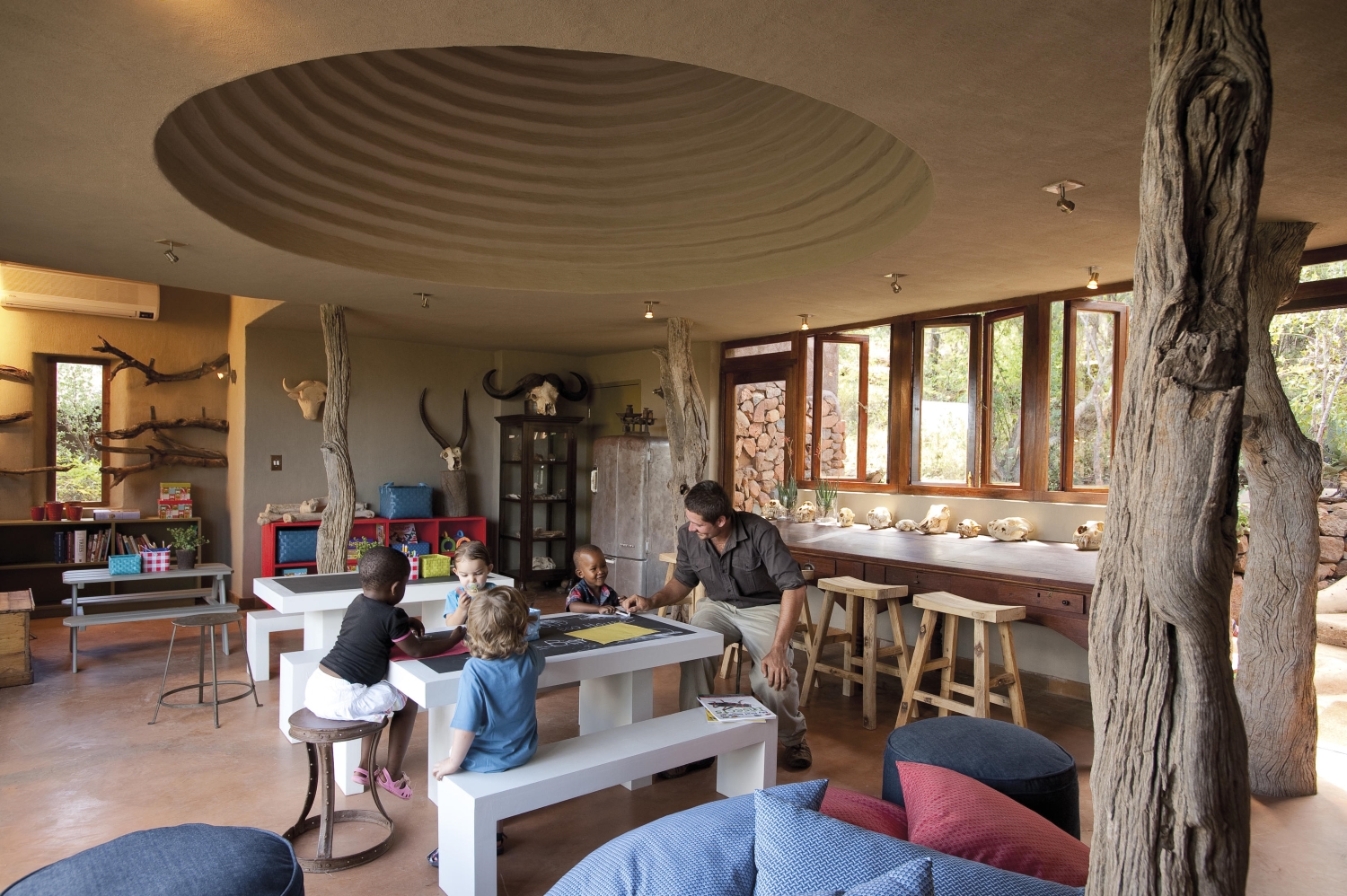 12-Madikwe-Safari-Lodge-malaria-free-safari-kidz-club An interactive kids' playroom with tables, stools, and a variety of educational items, where children engage in activities alongside a guide, surrounded by decor inspired by the African bush.