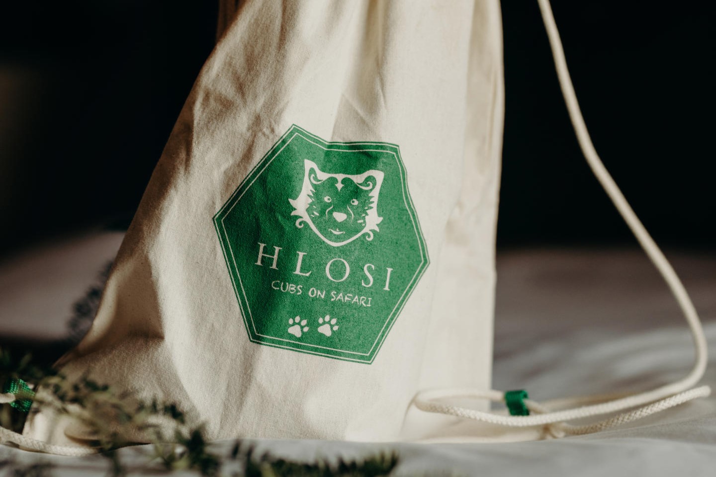 4-Amakhala-Hlosi-Game-Lodge-cubs-on-safari A drawstring bag with the green logo of "Hlosi Cubs on Safari," featuring a lion's face and paw prints, is placed on a soft surface with subtle foliage in the foreground.