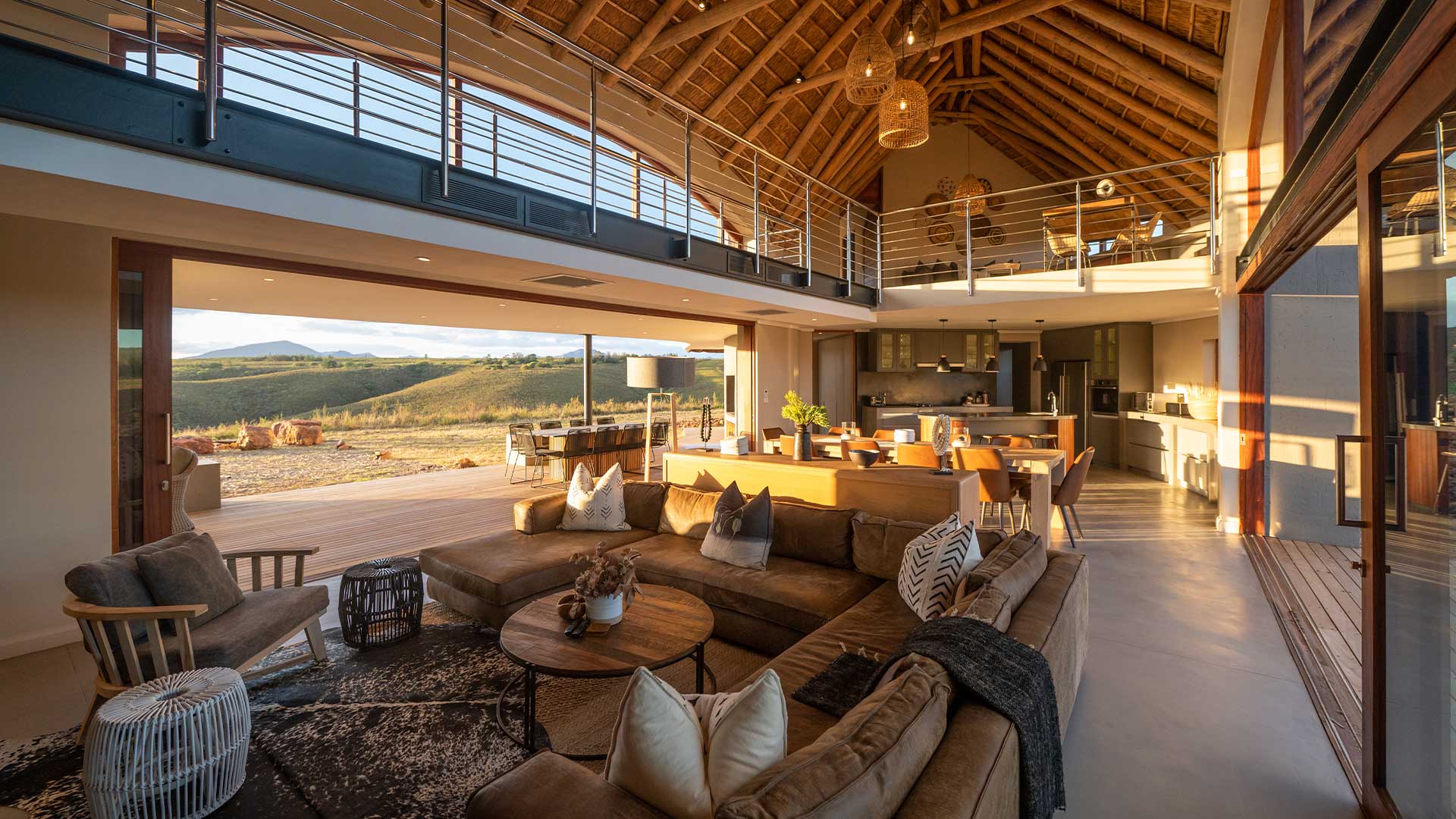 5-Gondwana-Kwena-villas A spacious, open-plan lodge interior with modern furnishings, a high thatched ceiling, and large glass doors offering a view of rolling hills bathed in golden sunlight.