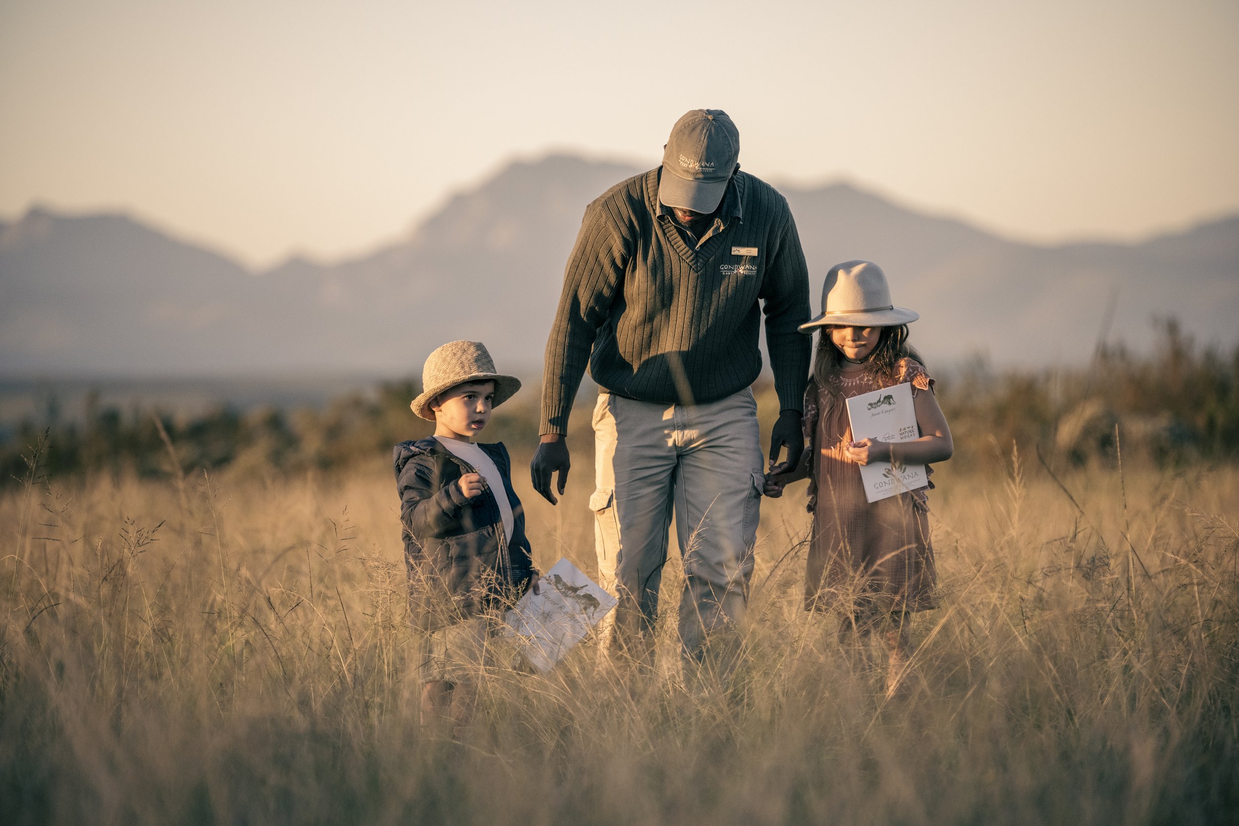 6-Gondwana-Junior-Rangers A ranger guides two young children through a grassy field at sunset, with mountains in the background. The children hold safari activity sheets and wear wide-brimmed hats.