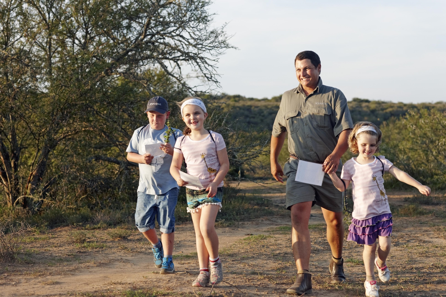 8-Kwandwe-Ecca-Lodge-malaria-free-safari-for-kids A smiling guide leads three children on a bush walk, with the kids eagerly holding activity sheets and looking engaged in their surroundings.