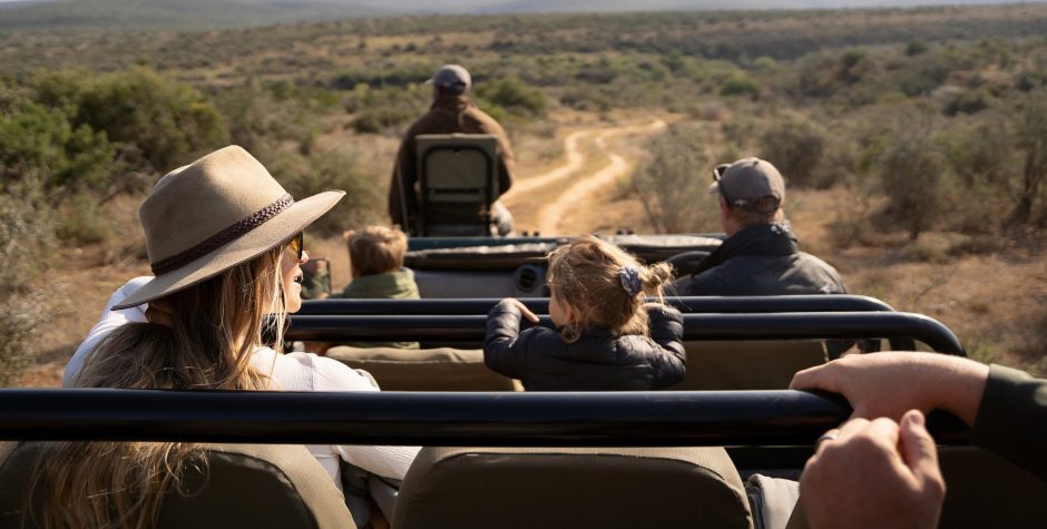 A family enjoys a safari game drive, seated in an open vehicle traversing a dirt road surrounded by vast bushveld, with a young child curiously looking out at the landscape.