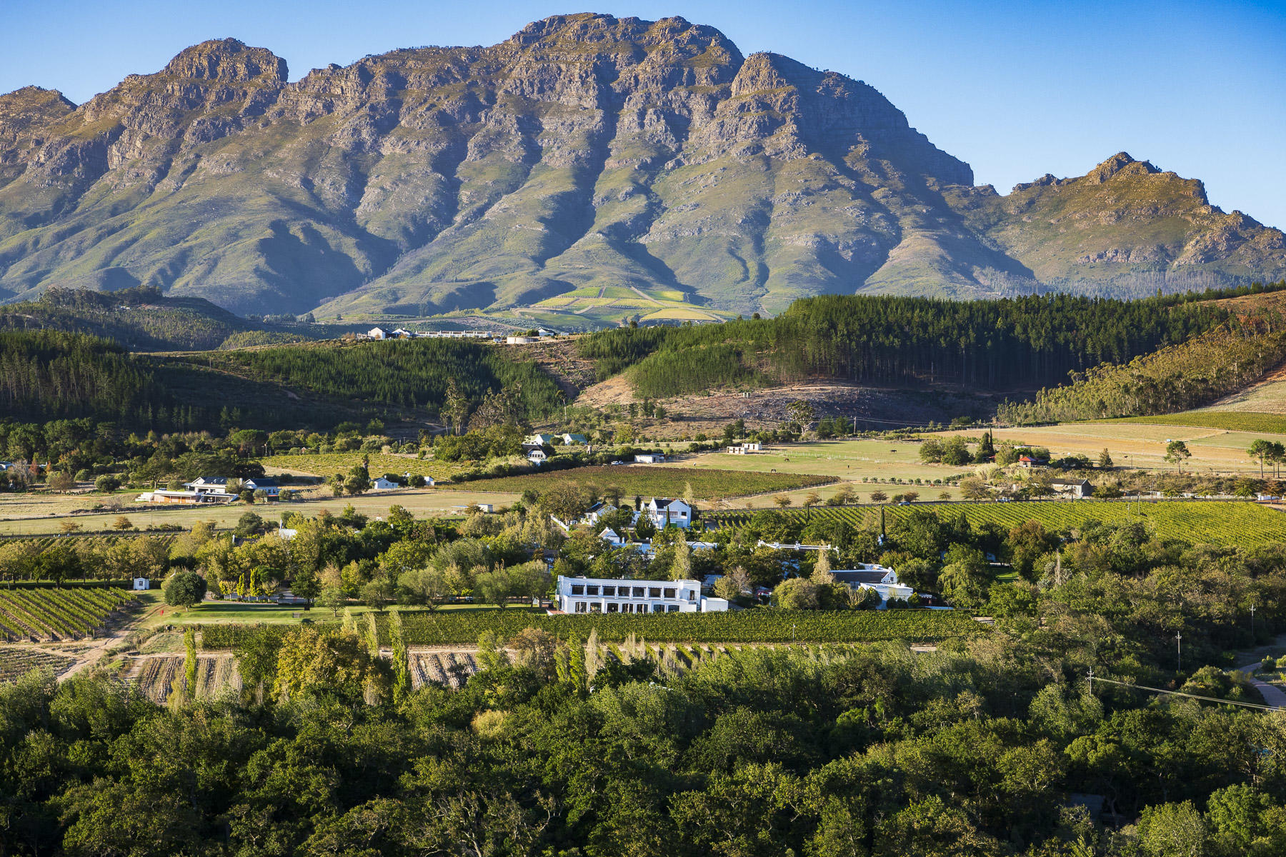 Lanzerac-Hotel-Spa A lush green valley with vineyards and white Cape Dutch buildings, framed by towering mountains under a clear blue sky