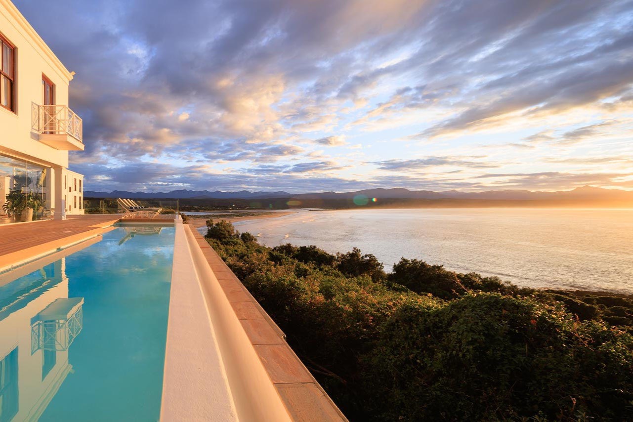 The-Plettenberg-Blue-Wing-Exterior A luxurious poolside view overlooking a serene beach and rolling hills, illuminated by the soft light of a sunset sky.