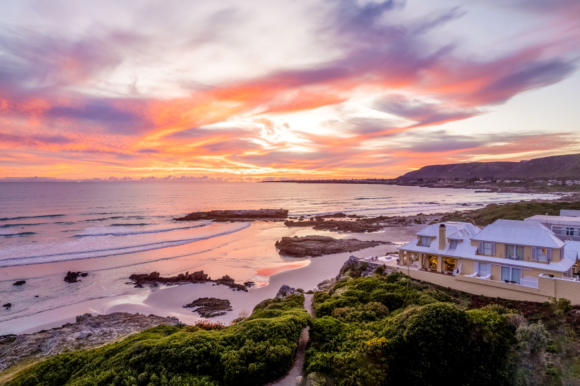 birkenhead_house_sunset A luxury coastal lodge overlooks the ocean at sunset, with vibrant pink and orange hues painting the sky