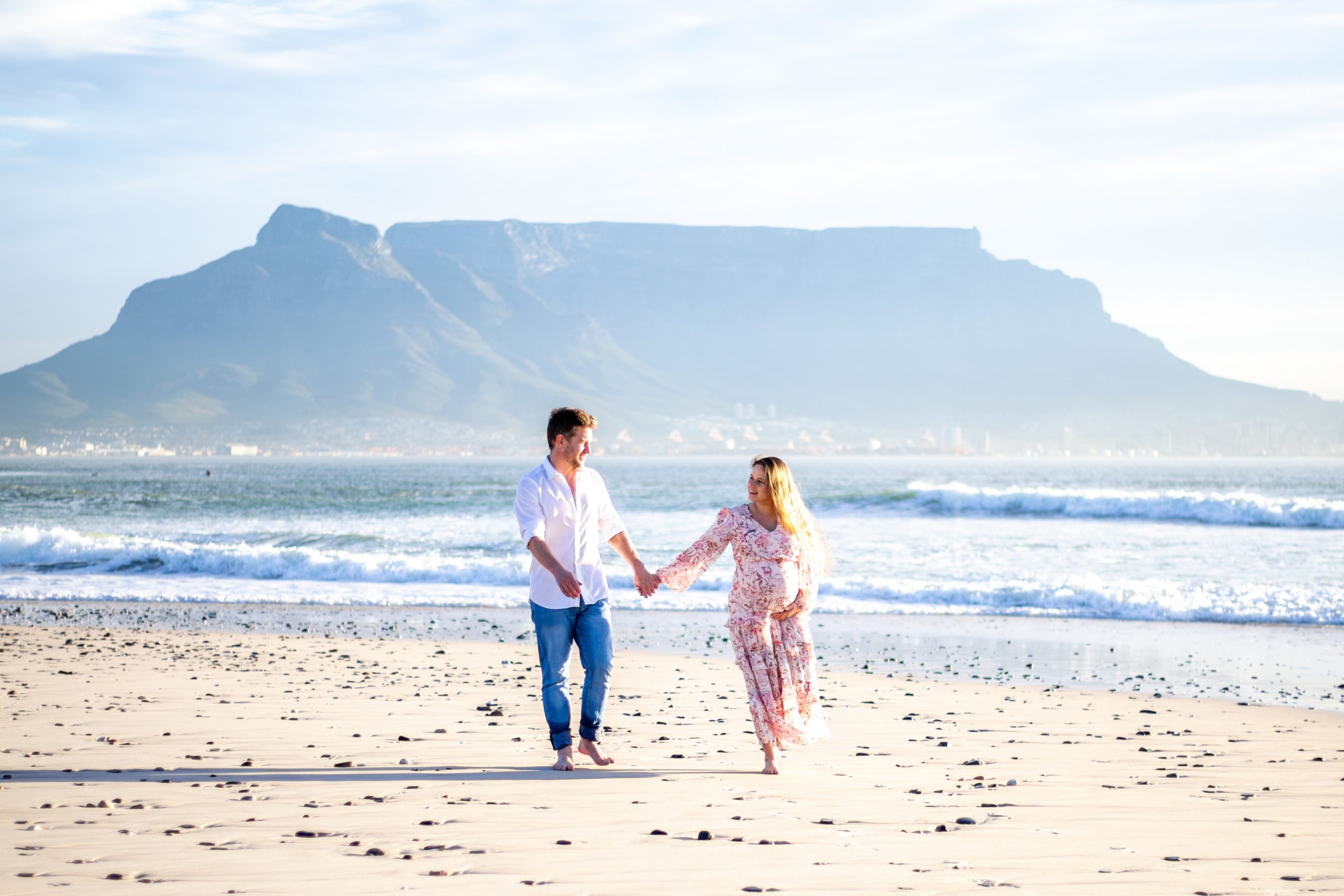 katharina-and-andrew-maternity-photos-sunset-beach-58 (1) Katharina in a flowing floral dress walks hand-in-hand with her husband along a sandy beach, with Table Mountain rising in the background during their babymoon safari.