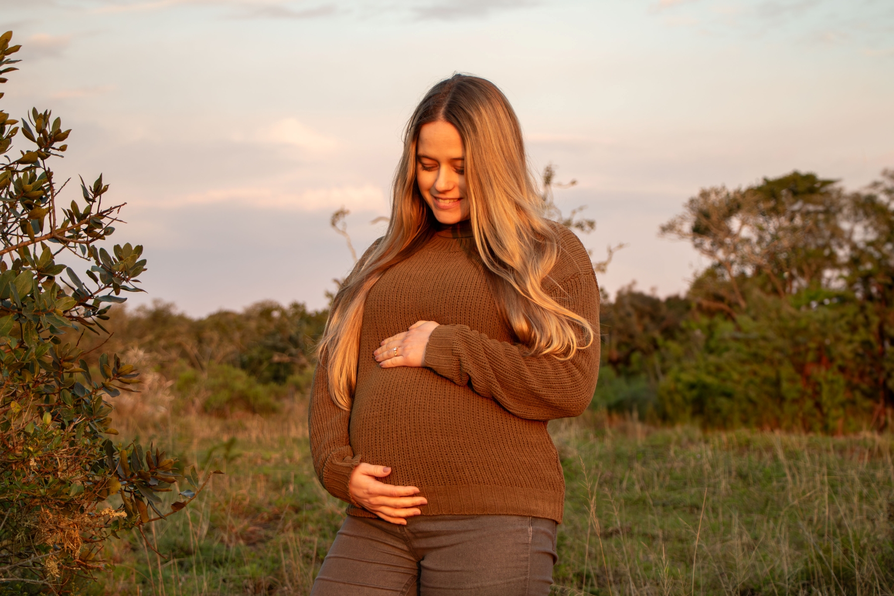 luxury-safaris-with-rhino-africa-at-amakhala_N8A9712 (1) stands in the African bush at sunset, smiling down at her baby bump with her hands gently cradling it during her babymoon safari.