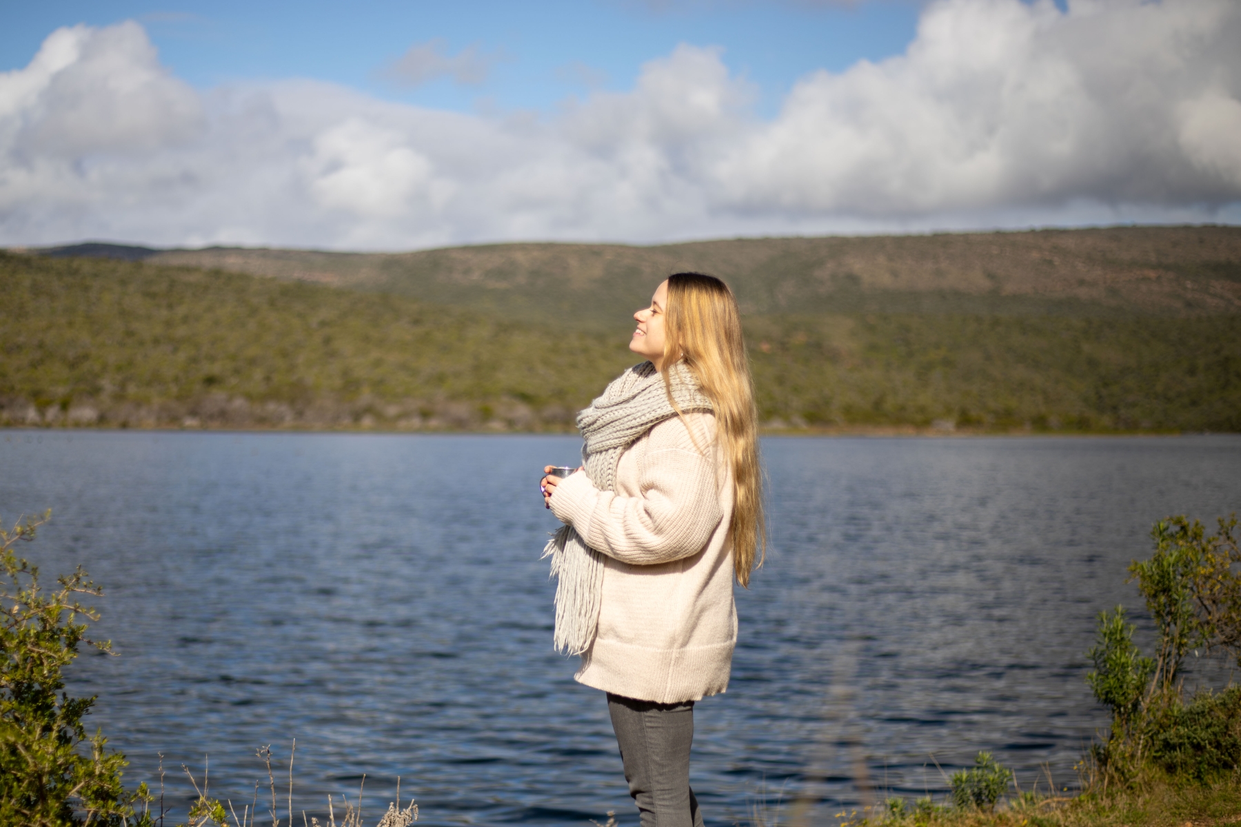 luxury-safaris-with-rhino-africa-at-kwandwe_D6A9959 Katharina stands by a tranquil lake, wrapped in a warm scarf, eyes closed as she tilts her face toward the sun during her babymoon safari.