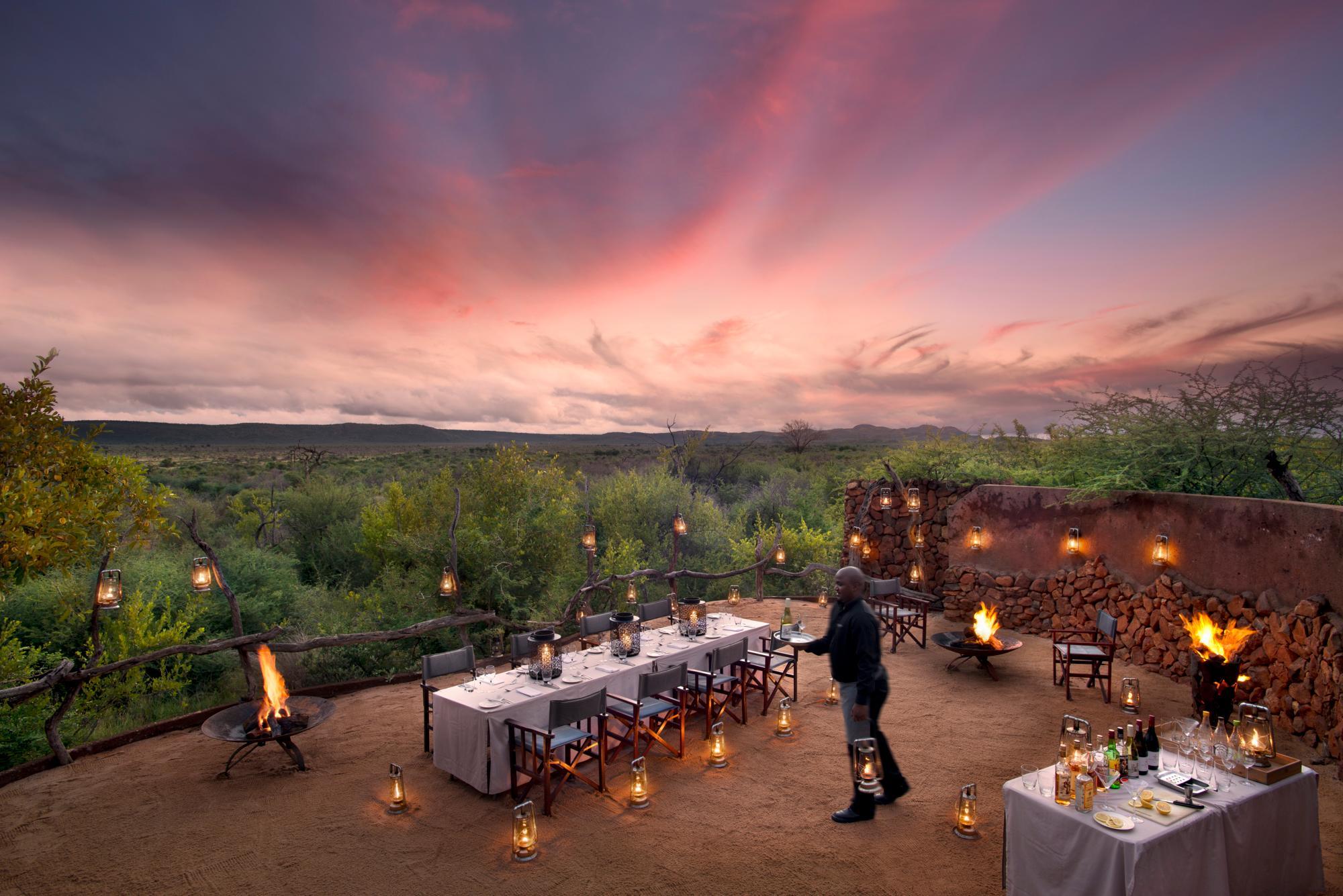 Madikwe Safari Lodge Kopano Lodge Boma dinner A beautifully set outdoor dining area, illuminated by lanterns and a central fire pit, overlooks a serene bush landscape at sunset, with a staff member preparing the tables.