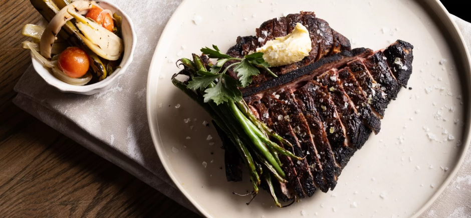 Marble-Cape-Town-restaurant-steak Top-down view of a sliced grilled bone-in steak topped with melting butter and served with charred green beans on a white plate, beside a small bowl of roasted vegetables.